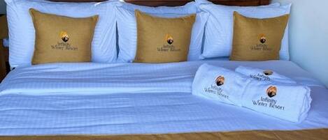 A neatly made bed with white and brown bedding, two pillows with a logo, and a folded towel with the same logo.
