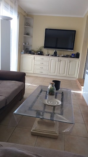 A living room with a sofa, a glass coffee table with decorative items, a flat-screen TV on the wall, and a white cabinet with drawers. - Le Mazet du bonheur Issambres (Roquebrune-sur-Argens)