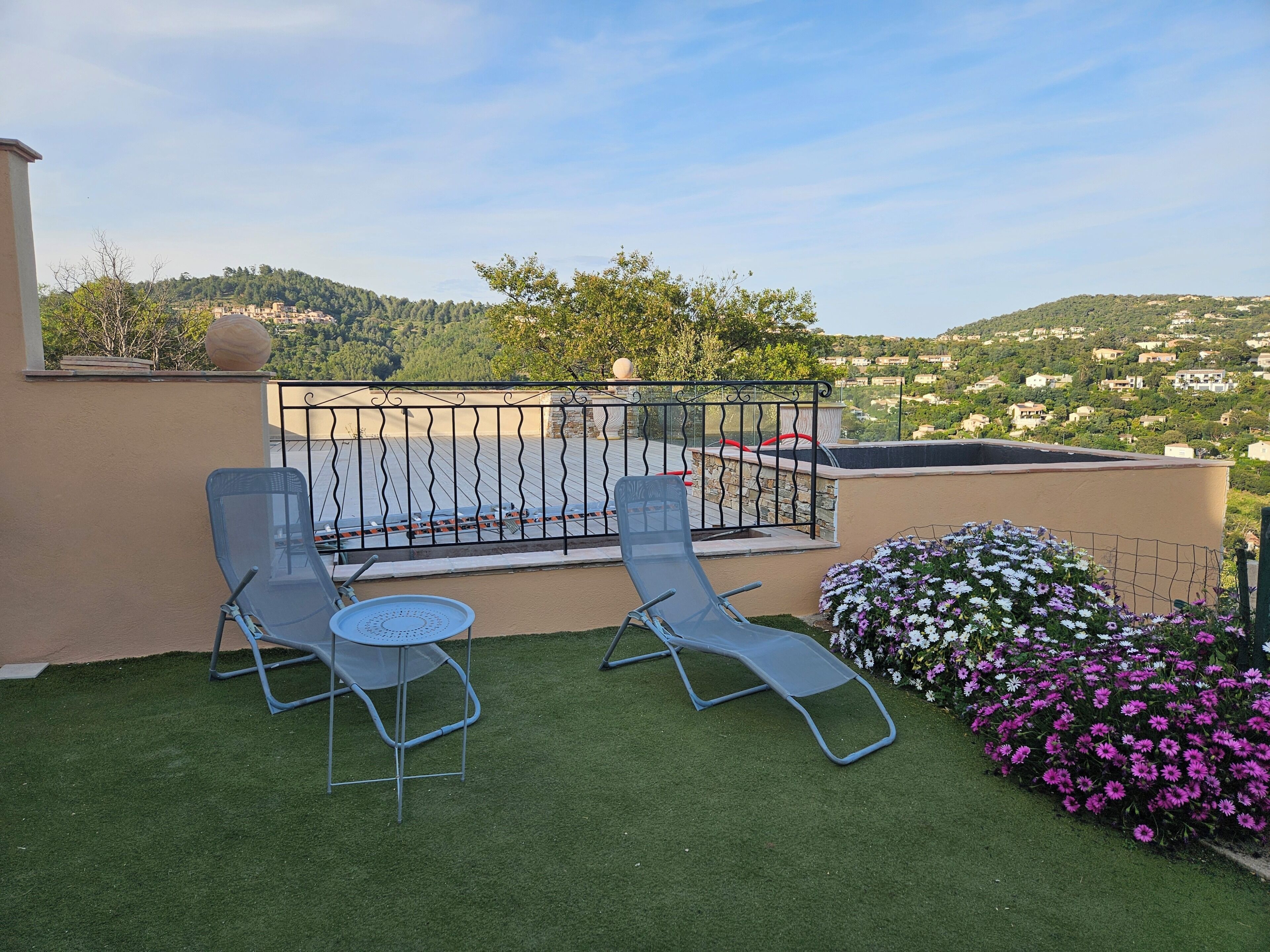 A balcony with two lounge chairs, a small table, and a flower bed with purple flowers.