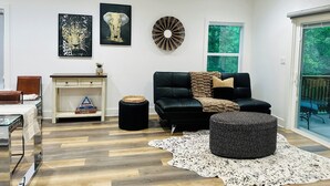 A modern living room with a black leather sofa, a glass coffee table, a decorative wall art piece, and a ceiling fan. - Deluxe Country Cabin - Pet Friendly  (Hot Springs)
