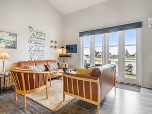 Una sala de estar moderna con sofá, sillón y mesa de centro, ventanas amplias y una pared con texto.