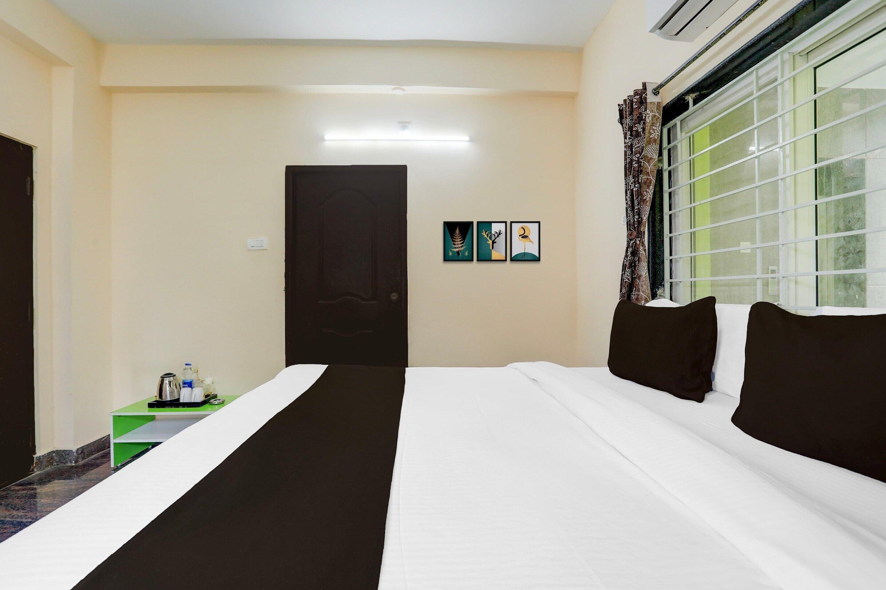 A hotel room with a bed, a small table with a kettle, a bottle, and a cup, a window with curtains, and a door.