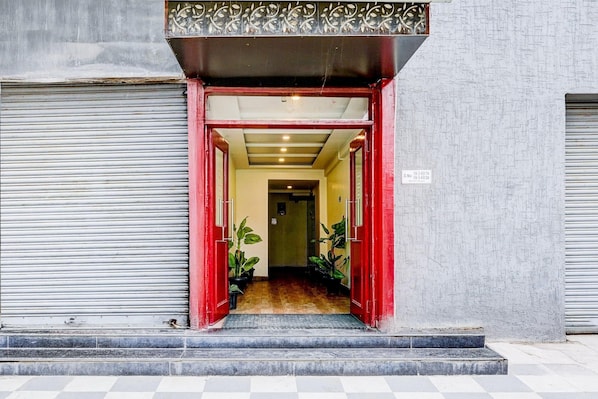 A building entrance with a red door, a metal shutter on the left, and a tiled floor.