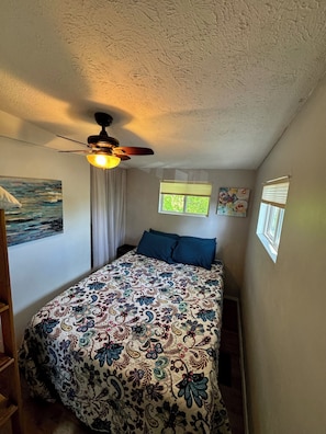 Ein Schlafzimmer mit einem Bett, einem Deckenventilator, zwei Fenstern mit Jalousien und einem Bild an der Wand.