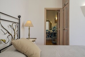 Ein Schlafzimmer mit einem Bett, einer Nachttischlampe, einem Wanddekor und Blick in ein Badezimmer mit Spiegel und Fliesen.