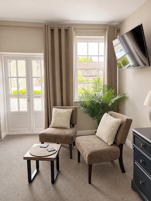 Ein Zimmer mit zwei Sesseln, einem kleinen Tisch, einem an der Wand befestigten Fernseher und einem großen Fenster mit Vorhängen.