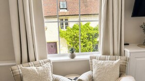 Ein Zimmer mit zwei Sesseln, einem kleinen Tisch, einem Fenster mit Vorhängen, einer Zimmerpflanze und einem Fernseher auf einem Schrank.