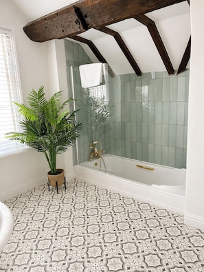 Ein Badezimmer mit Badewanne, Dusche, gemusterten Bodenfliesen, einer Topfpflanze und einem Fenster mit Jalousien.
