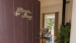 En trædør med 'The Coach House' skilt, der fører ind i et rum med trægulv, en stol, et bord og en potteplante.