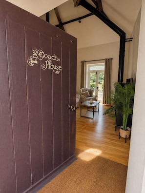 A wooden door with 'The Coach House' written on it, leading into a room with a wooden floor, a chair, a table, and a potted plant. - The Coach House | Central Location (Shropshire)