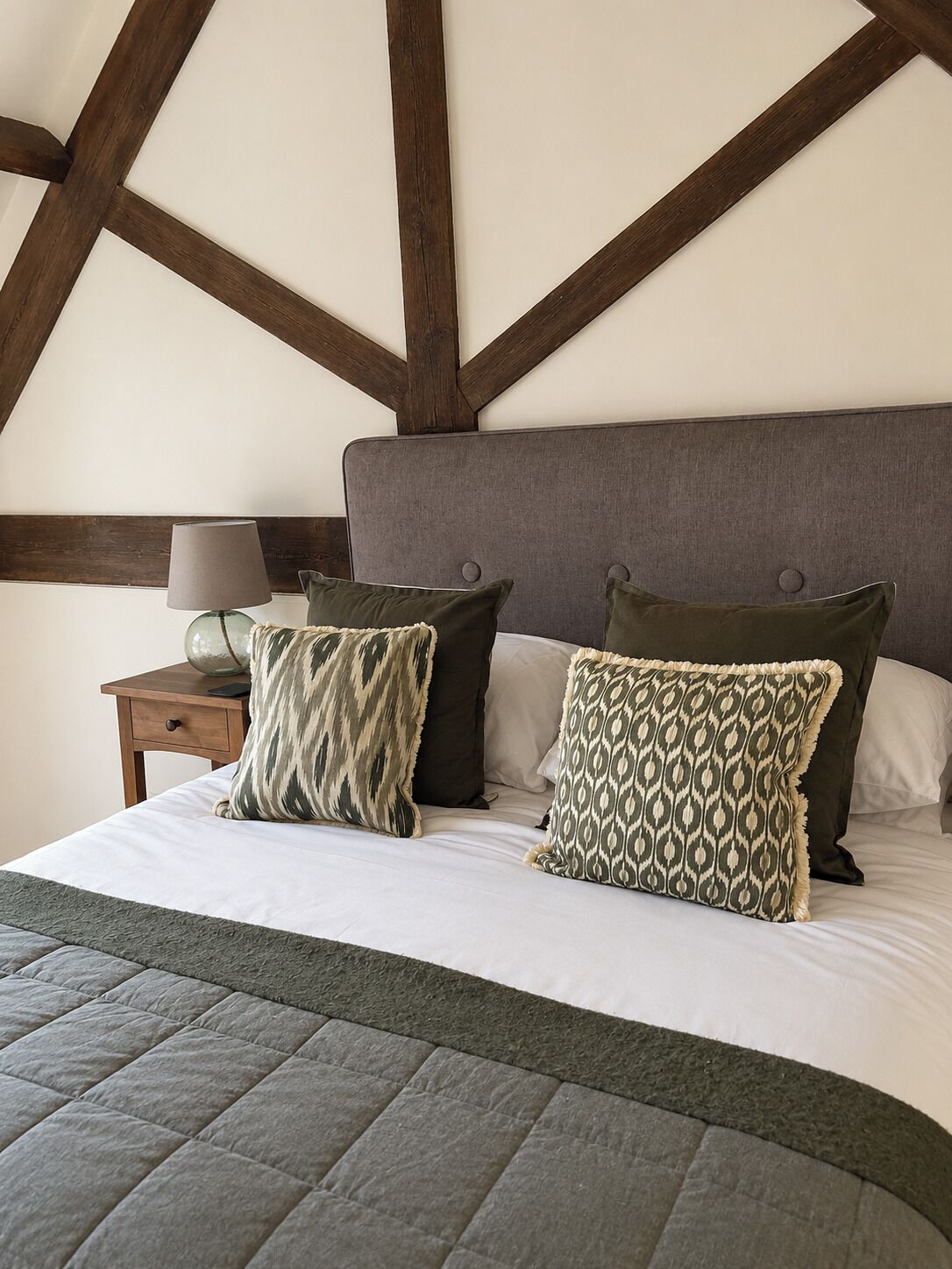 A bedroom with a bed, pillows, a nightstand, a lamp, and exposed wooden beams on the ceiling.