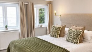 Een slaapkamer met een groot bed, beige gordijnen, een bedlampje en uitzicht op groen.