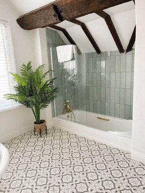 Ein Badezimmer mit Badewanne, Dusche, gemusterten Bodenfliesen, einer Topfpflanze und einem Fenster mit Jalousien.