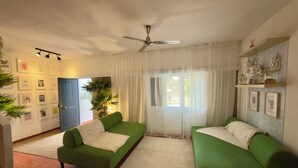 Un salon avec deux canapés verts, un ventilateur de plafond, une grande fenêtre avec des rideaux blancs et un mur décoré de tableaux encadrés.
