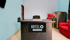 Un comptoir d’accueil portant l’inscription « HOTELO », agrémenté d’un panneau indiquant qu’il est mis à disposition par l’entreprise, ainsi qu’une chaise rouge, un réfrigérateur et un pot de fleurs.