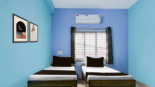 Une chambre avec deux lits en bois, une fenêtre avec des rideaux, un climatiseur et deux tableaux encadrés accrochés au mur.