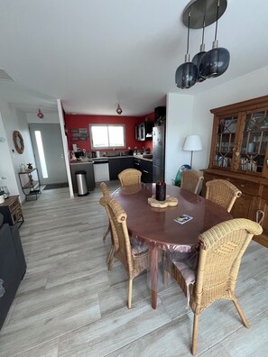 A dining area with a round table, wicker chairs, a modern kitchen in the background, and a cabinet with glass doors. - Quiet new house 2.5 km from the sea, the Zoo and the resort of La Palmyre (Saint-Augustin)