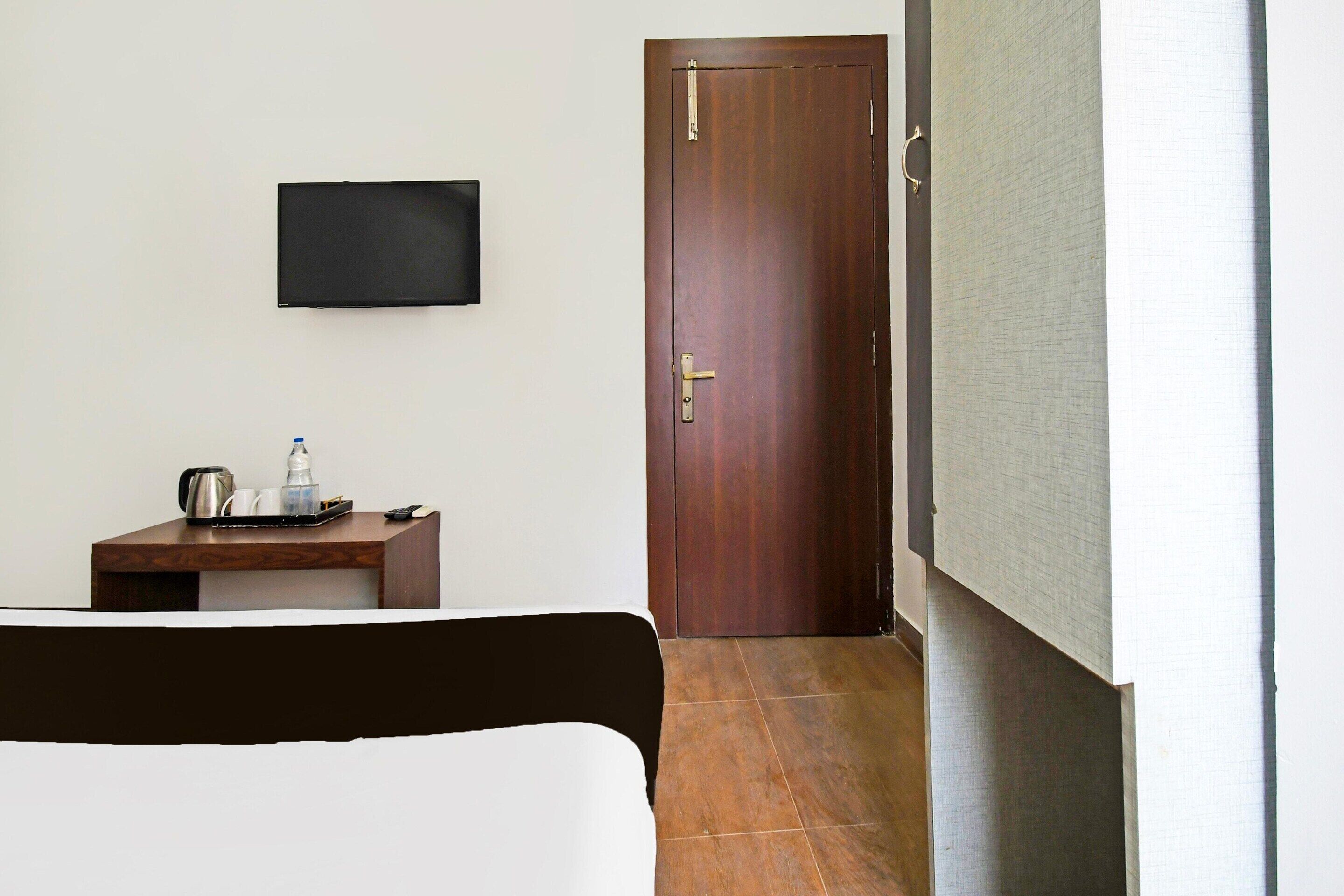 A hotel room with a bed, a desk with a kettle, a bottle, and a remote control. There is a door and a wall-mounted TV.