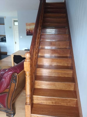 Un escalier en bois avec une rampe, menant à une pièce où se trouvent une table à manger et un canapé en cuir rouge.