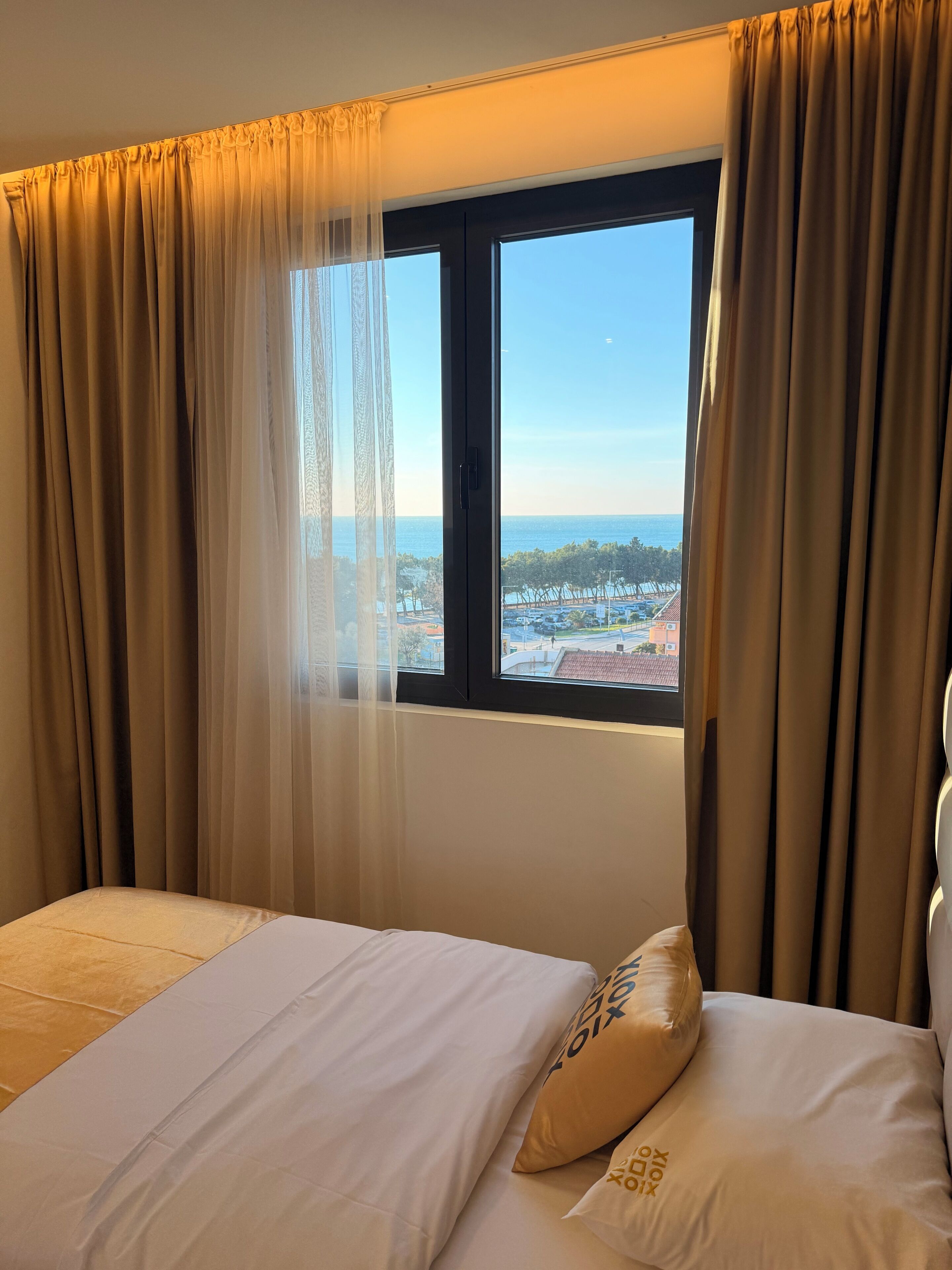 A hotel room with a bed, a window with a view of the sea, and curtains.
