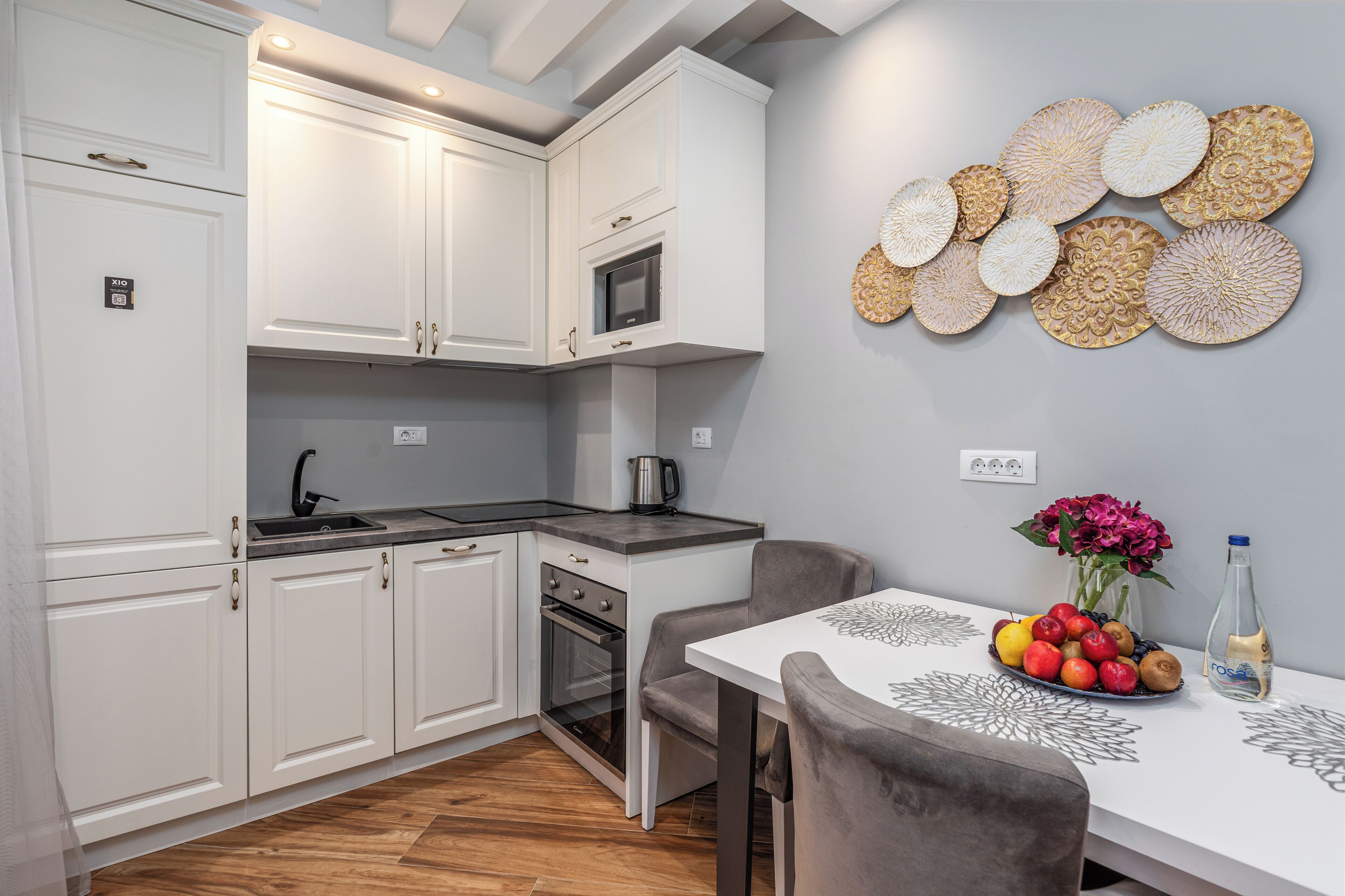 A modern kitchen with white cabinets, a microwave, and a sink. There is a dining table with a vase of flowers and a bottle of water.