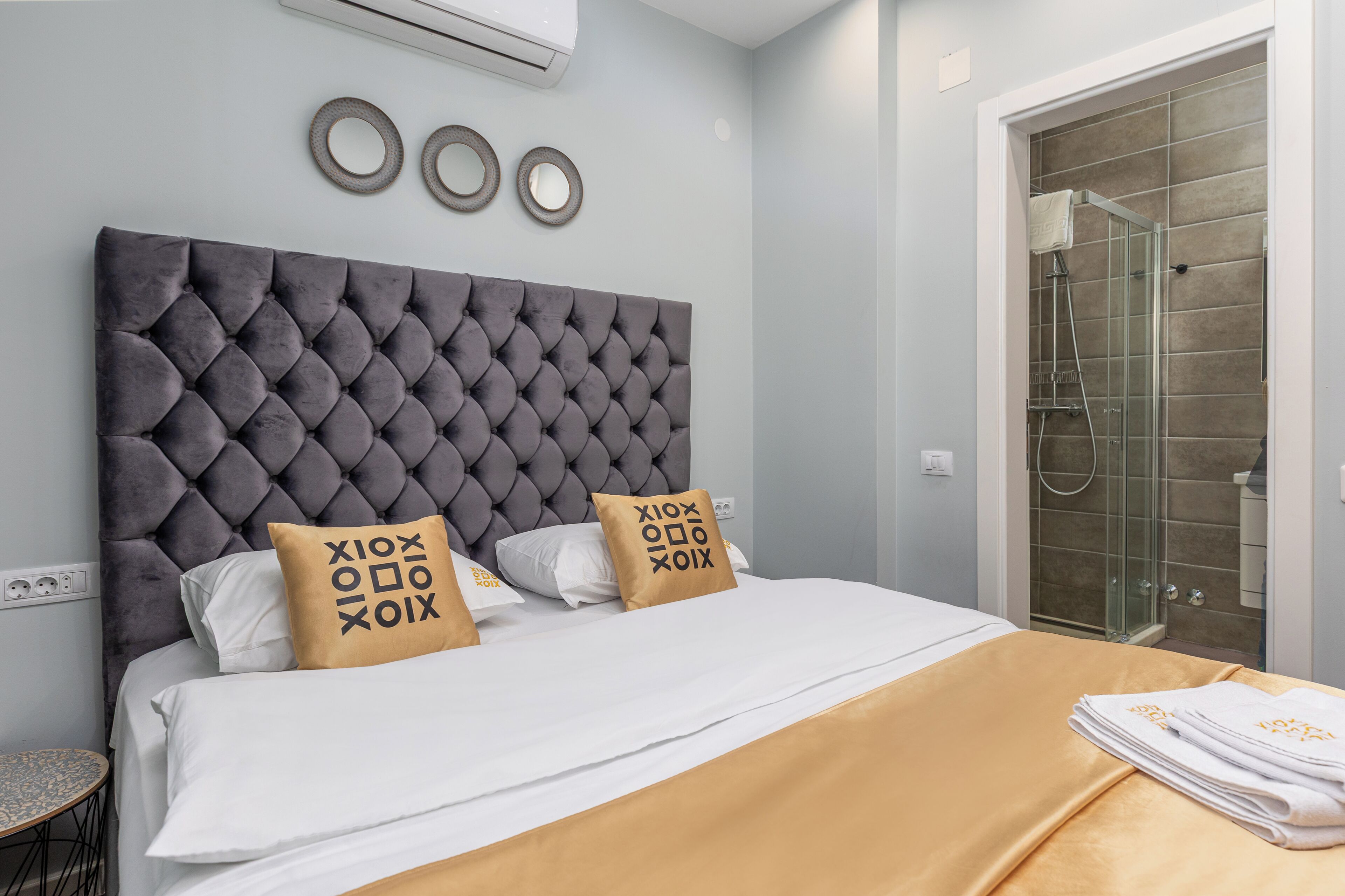 A bedroom with a tufted headboard, a bed with white and gold bedding, and a glass shower enclosure.