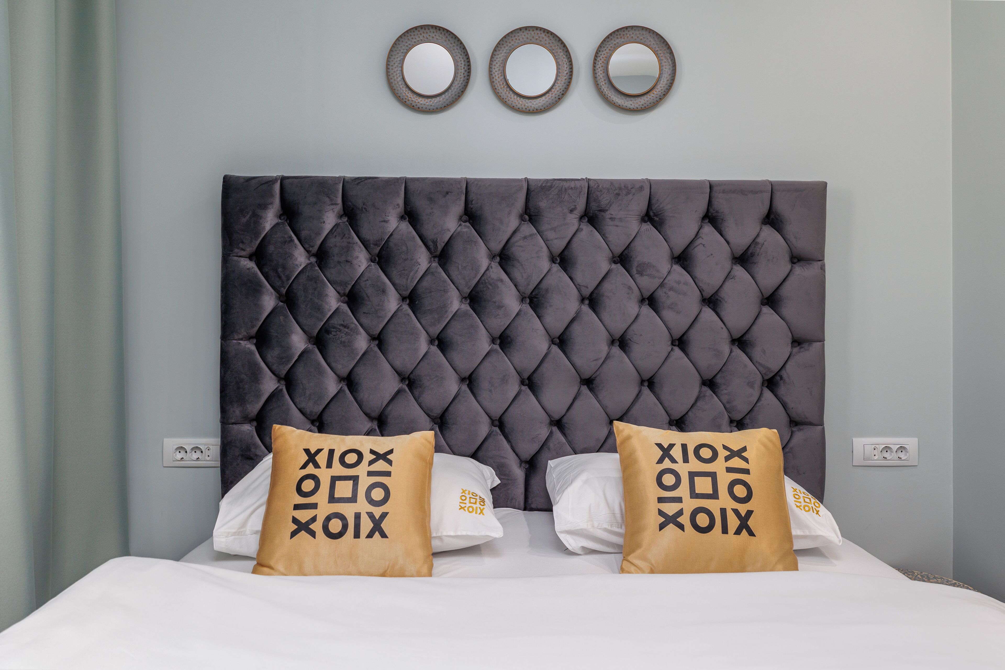 Een bed met een gestoffeerd hoofdbord, twee kussens met een logo en drie spiegels aan de muur.