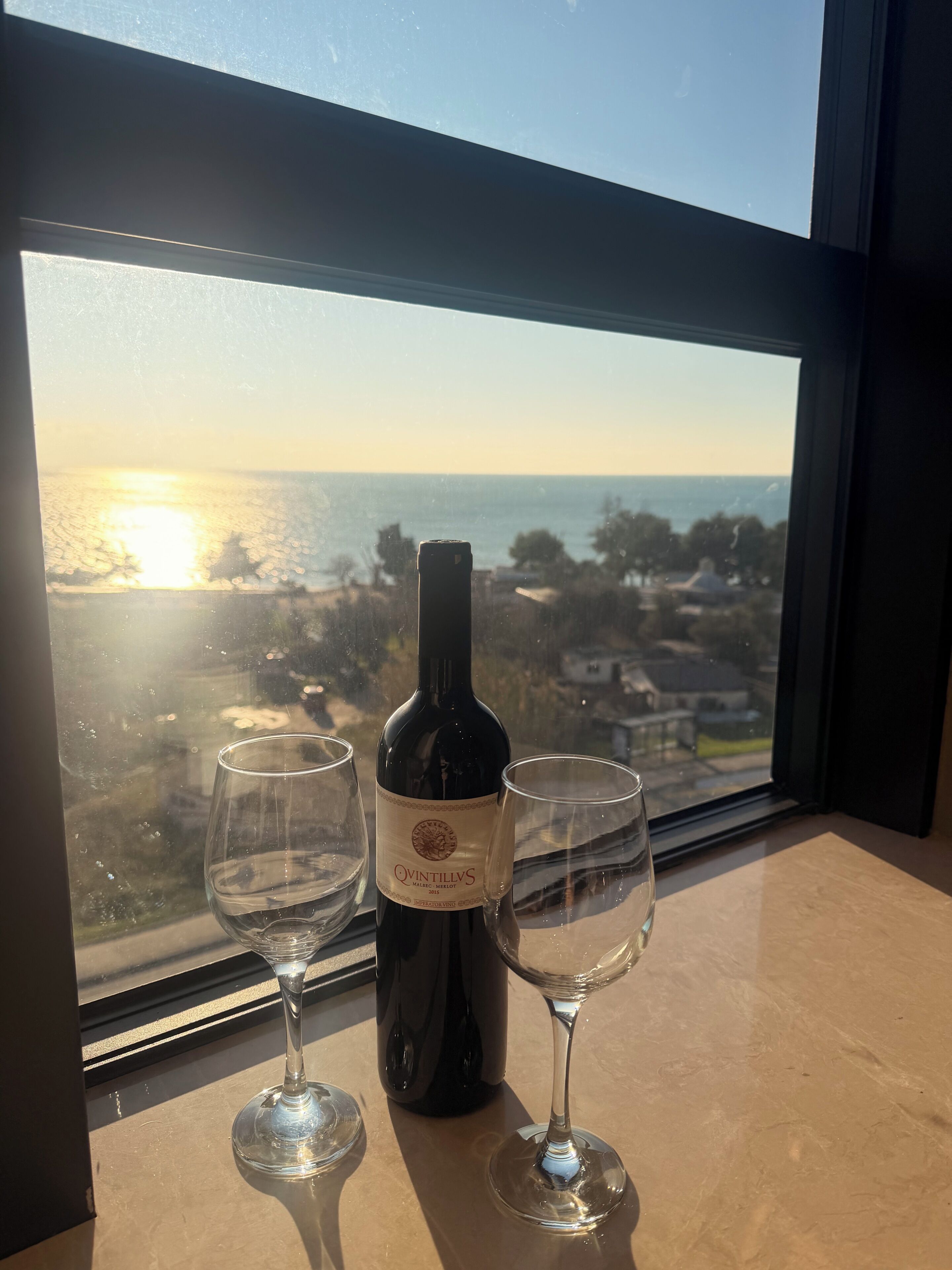 A wine bottle and two glasses on a table by a window with a view of the ocean.