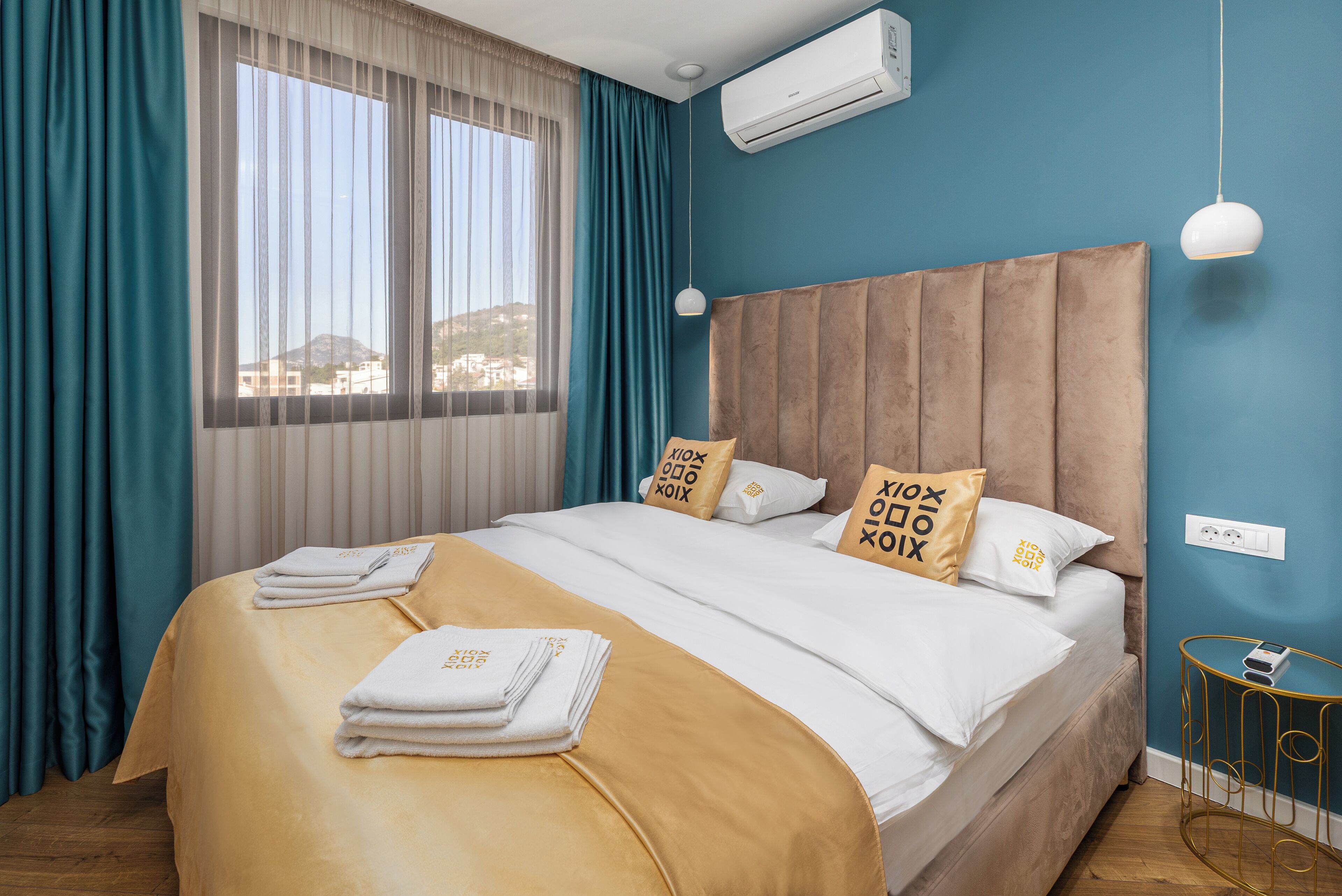 A hotel room with a bed, curtains, an air conditioning unit, a bedside table, and a view of buildings.