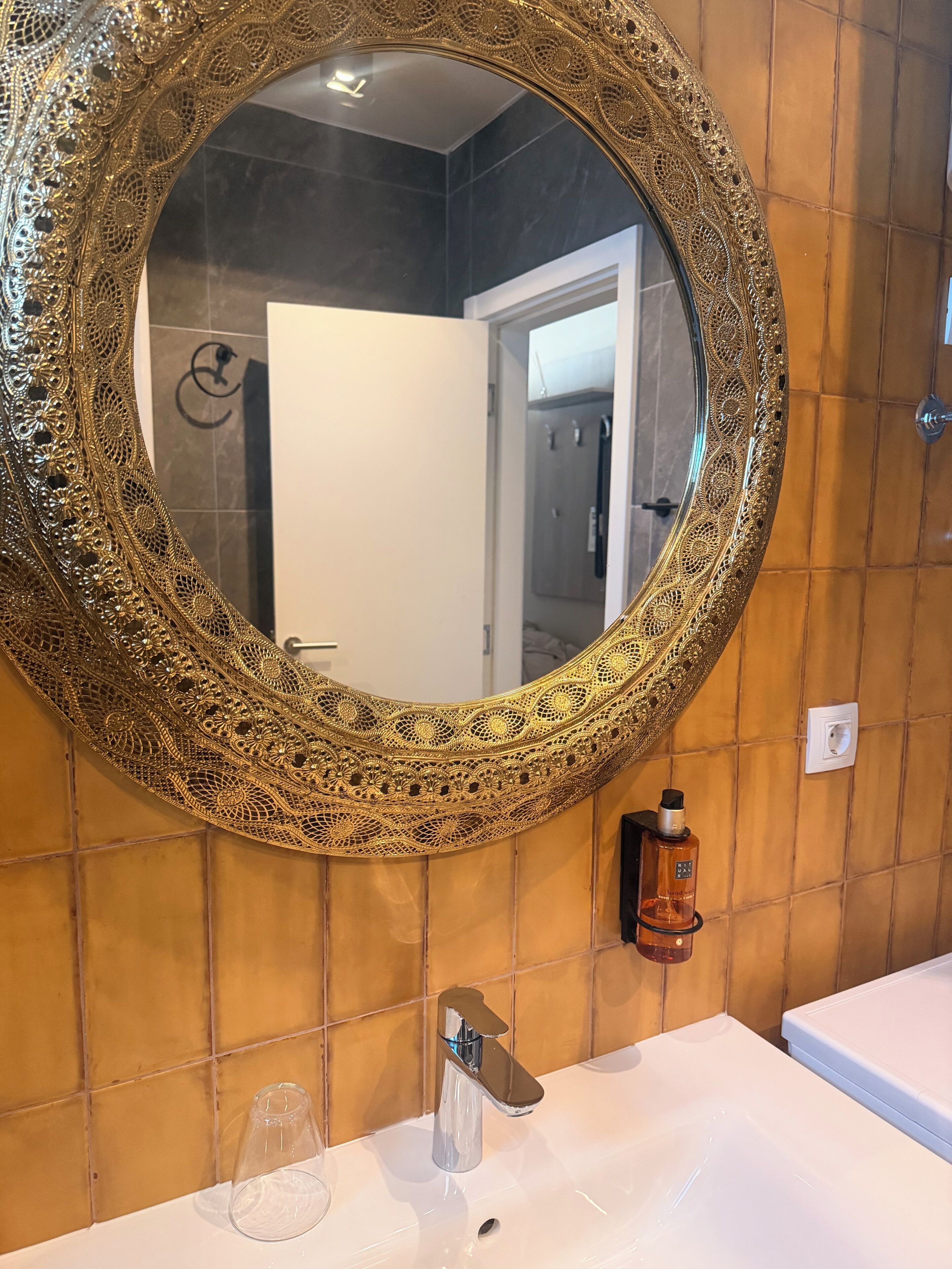A bathroom with a round ornate mirror, a white sink, and a bottle of hand soap.