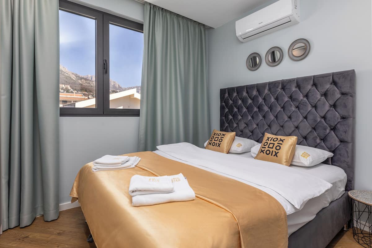 A hotel room with a bed, a window, curtains, an air conditioning unit, and a wall-mounted mirror.