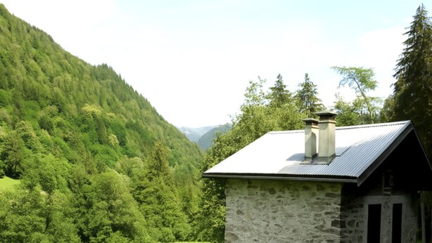 A stone building with a metal roof, surrounded by lush green fields and forested hills.