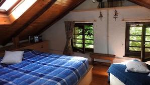 A bedroom with a sloped ceiling, a bed with a blue and white checkered bedspread, a wooden bench, and a window with a view of greenery.