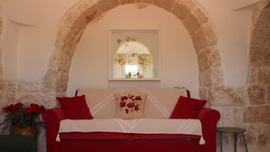 A red sofa with a floral pillow, a small table, and a vase with red flowers against a stone wall with an arched window. - Cozy medieval trullo for couples and families seeking tranquility. (OSTUNI)