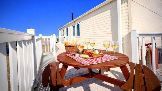 Una mesa de picnic de madera con mantel a cuadros, rodeada de sillas de madera, ubicada en una terraza con valla blanca.