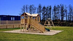 Una estructura de juego de madera con un tobogán y una escalera de cuerda, rodeada por una cerca y un edificio al fondo.