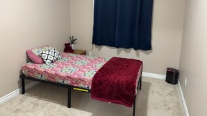 A small bedroom with a single bed, a nightstand, a red teddy bear, a potted plant, and a dark curtain.
