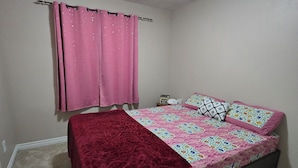 A bedroom with a bed, a window with pink curtains, a ceiling light, and a carpeted floor.