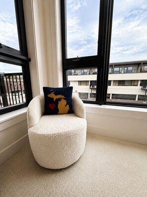 Un sillón redondo y mullido con un cojín que presenta un diseño de mapa y corazón, situado junto a una ventana con vistas a edificios.