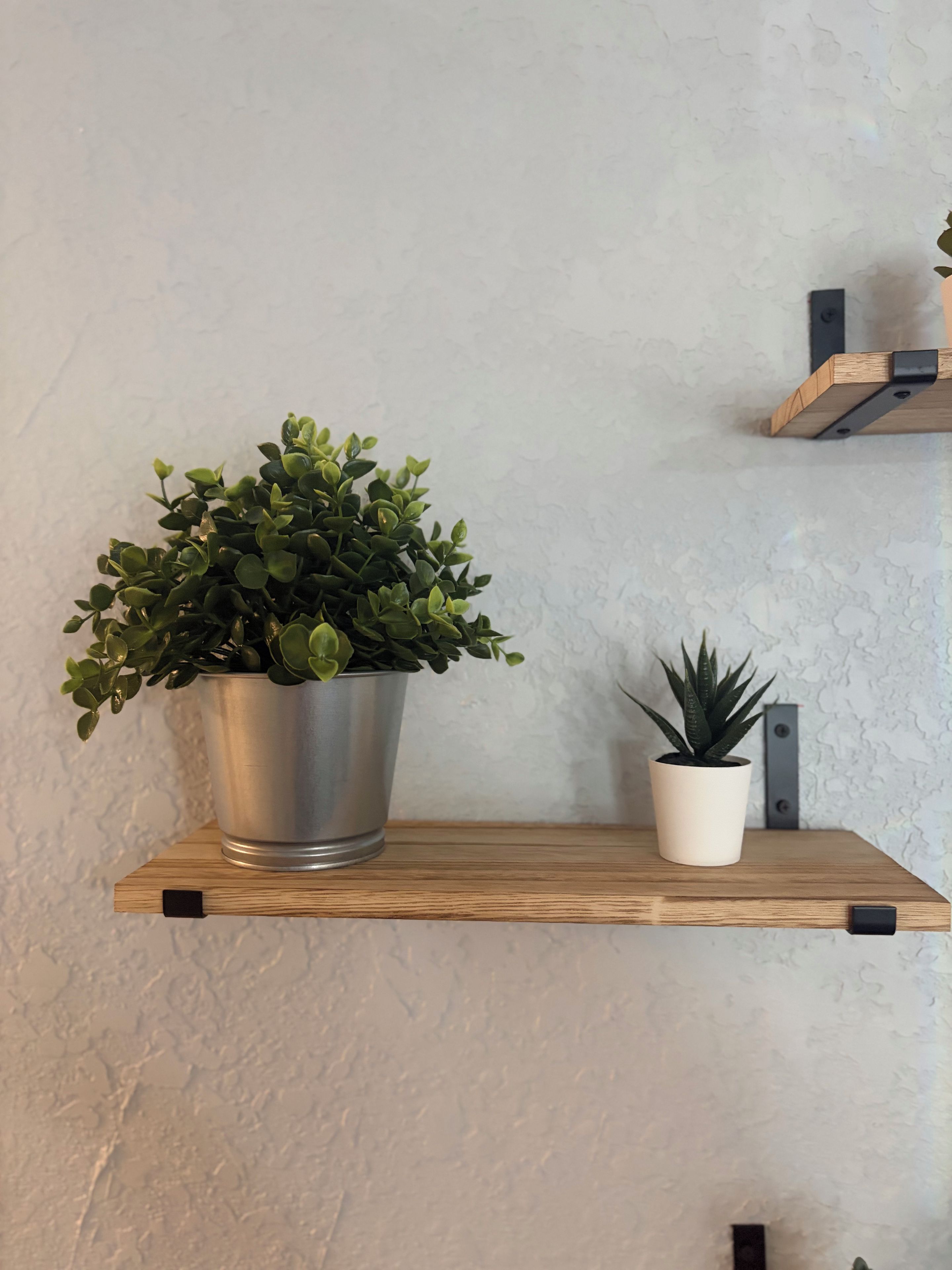 A shelf with two potted plants, one in a metal pot and one in a white pot, against a textured wall.