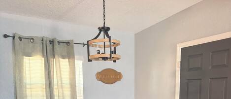 A dining area with a dark wooden table, four chairs with yellow upholstery, a hanging light fixture, and a 'WELCOME' sign on the wall.