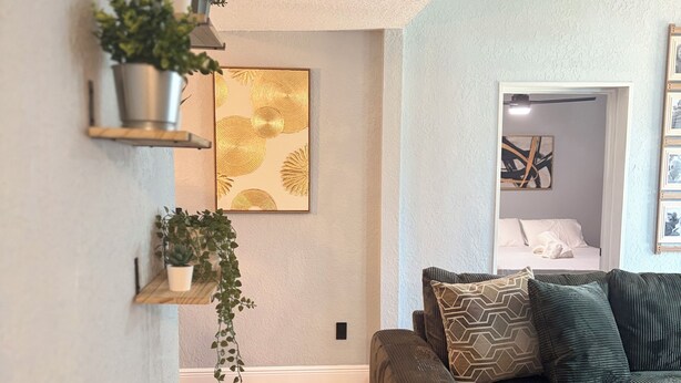 A living room with a wooden floor, a green sofa, and a wall-mounted shelf with plants. - Amelias Arena Retreat 4bedroom home in Orlando (Orlando)