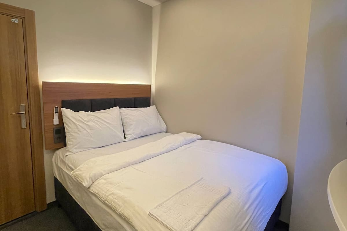 A hotel room with a bed, a wooden door, a bedside table with a lamp, and a bathroom with a bathtub.