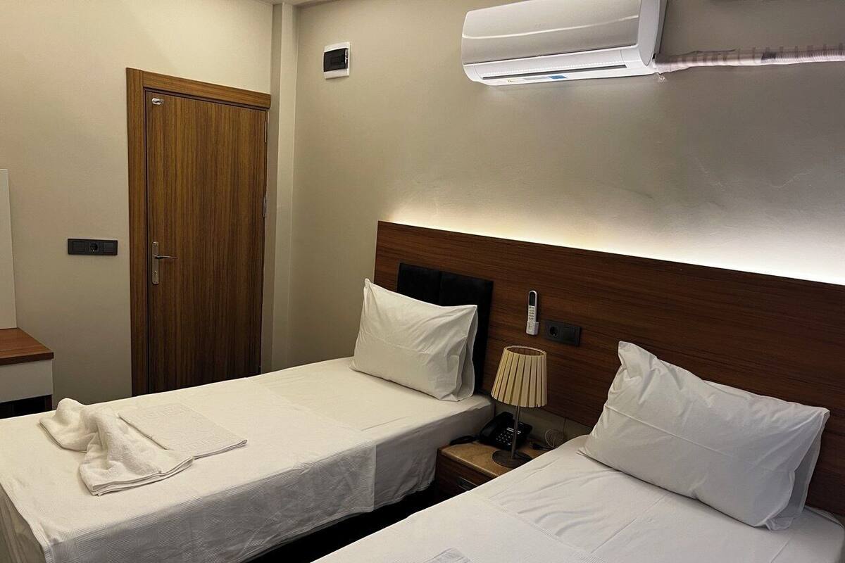 A hotel room with two beds, a wooden door, a bedside table with a lamp, and an air conditioning unit.