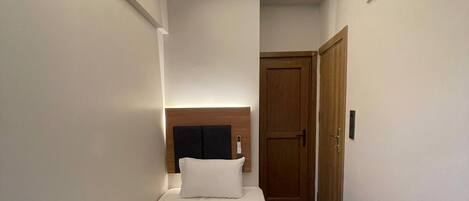 A single bed with a headboard, a bedside table, a wooden door, and a small wall-mounted light fixture.