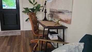 Una habitación con suelo de madera, un sofá verde, un escritorio con lámpara, una silla, una planta en maceta y un cuadro en la pared.