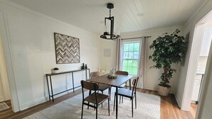 Un coin repas comprenant une table en bois, des chaises, un tapis, une suspension, une plante en pot et une fenêtre avec des rideaux.