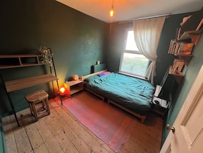 A room with a bed, a small stool, a shelf with books, a window with curtains, and a hanging light bulb. - Room-cozy Guest Room in Vibrant Brighton (Brighton)