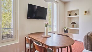 Un coin repas avec une table en bois, des chaises, un vase de fleurs, un téléviseur à écran plat et une fenêtre donnant sur des arbres.