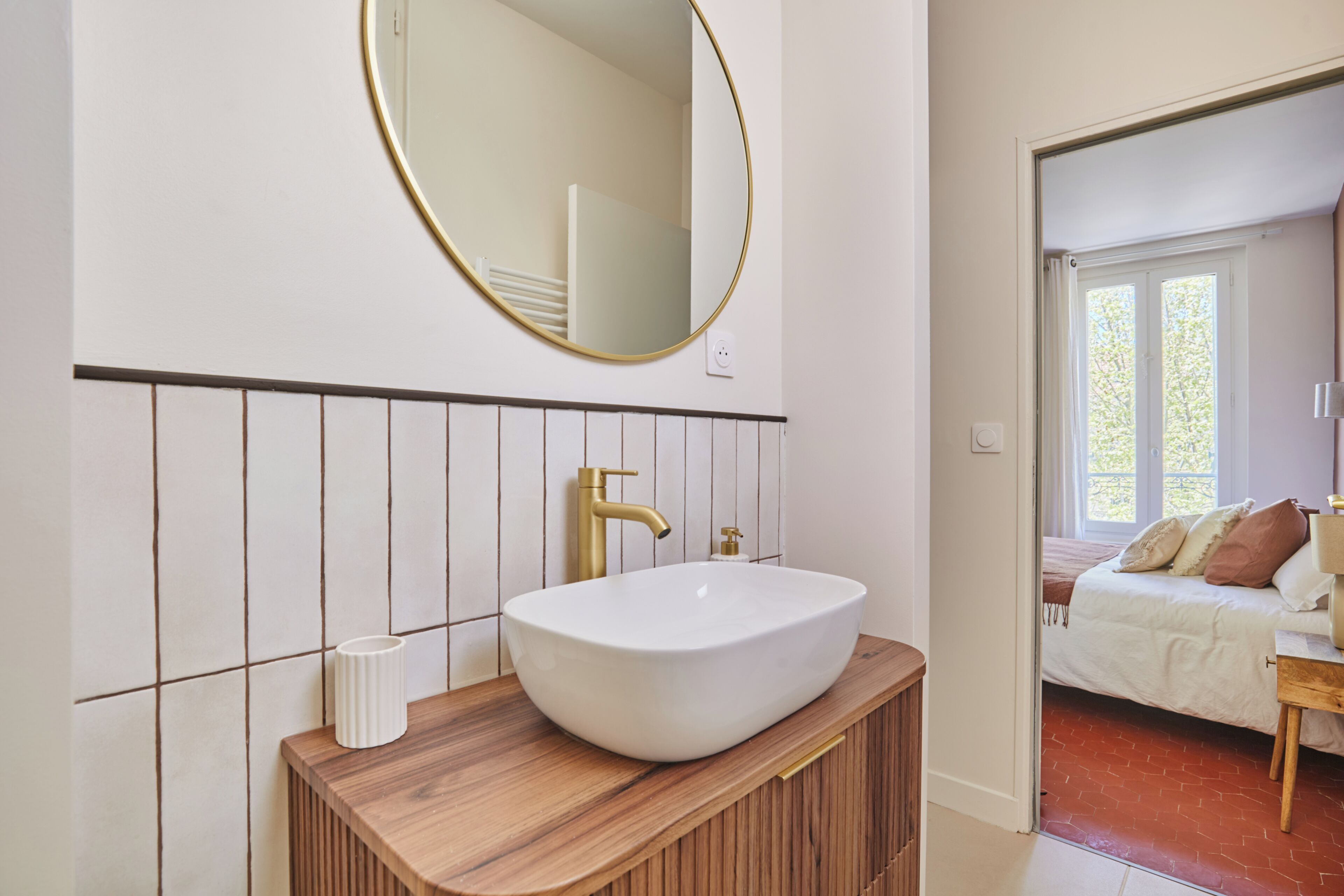 Un baño con lavamanos blanco, grifo dorado y espejo redondo sobre un mueble de madera.